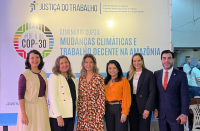Foto de cinco mulheres e um homem de pé e sorrindo. Atrás deles, painel de cor clara com informações sobre o evento.