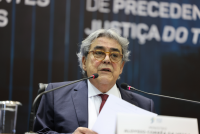 Foto de um homem claro, de cabelos grisalhos e usando óculos. Ele fala ao microfone enquanto segura papéis que lê.