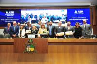 Foto mostra um grupo formado por homens e mulheres, em pé, atrás de uma bancada. Ao fundo, um painel eletrônico ligado com o destaque para a logomarca da ALEMA - Assembléia Legislativa do Maranhão