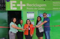 Foto mostra cinco pessoas em pé, fazendo o corte de uma fita inaugural. Ao fundo um painel verde com informações em letras brancas: E + RECICLAGEM. Posto de Coleta.