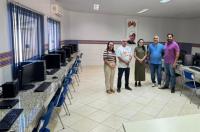 Foto mostra cinco pessoas em pé no fundo de uma sala comprida. Nas laterias, vários computadores instalados sobre as mesas.