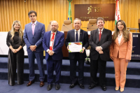 Foto de quatro homens ladeados por duas mulheres. Todos estão de pé e sorrindo para a foto. Um dos homens segura um certificado de premiação. Atrás deles, parede de madeira ripada e placa marrom com a inscrição TST em dourado.