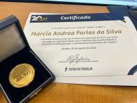 Foto mostra uma medalha e um certificado com destaque para o nome no documento: MÁRCIA ANDREA FARIAS DA SILVA