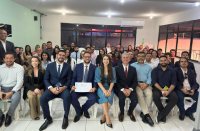 Foto mostra várias pessoas sentadas em plateia, olhando e sorrindo para a câmera. Atrás delas, uma parede verde e duas brancas, onde se localizam janelas de vidro.