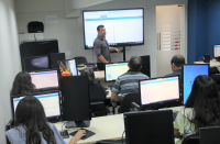 Foto de um homem claro, usando camisa cinza e dando aula para uma turma que usa computadores. Atrás dele, tela de projeção com apresentação de slides. O ambiente é uma sala de treinamento de informática com paredes brancas e azuis.