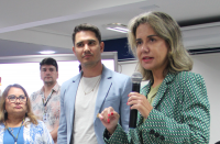 Foto de duas mulheres e dois homens. Em primeiro plano, à direita, mulher clara, de cabelos claros, usando blazer verde, fala ao microfone, que segura com a mão esquerda, e gesticula com a mão direita, olhando para a frente. Ao lado dela, as demais pessoas a observam falar.