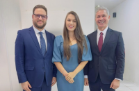 Foto mostra uma mulher clara, de cabelos castanhos compridos e usando vestido azul claro, ladeada por dois homens claros e usando ternos azuis escuros. Atrás deles, parede branca.