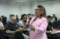 Foto de mulher clara, usando óculos e blazer rosa. Ela fala ao microfone com a mão direita, dirigindo-se à plateia de várias pessoas em ambiente de auditório com paredes brancas.