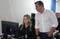 Foto de um homem claro, usando camisa branca e observando a tela de um computador. Ele está de pé ao lado de uma mulher clara de cabelos claros e compridos, usando uma blusa preta e utilizando o computador.