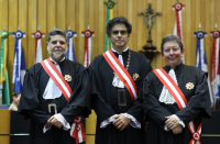 Foto mostra três homens perfilados um ao lado do outro. Ambos usam trajes formais. Ao fundo estão posicionadas as bandeiras dos estados brasileiros.