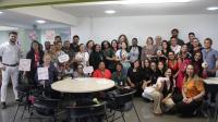 Foto mostra um grupo de pessoas, entre homens e mulheres reunidos em pé, e algumas pessoas agachadas. Alguns seguram nas mãos placas educativas sobre os cuidados contra o câncer de mama