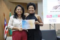 Foto de duas mulheres que estão em pé, em um auditório. Elas seguram um certificado.