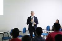 Foto mostra um homem branco usando um terno escuro sem gravata em pé, em uma sala de aula. Algumas pessoas estão sentadas.