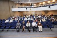 Foto mostra pessoas sentadas em cadeiras dentro de um auditório. Ao fundo, é possivel avistar um mesanino.