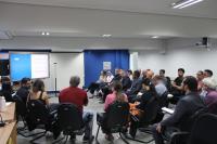 Foto mostra um grupo de pessoas. Todos estão sentados. E a distribuição das cadeiras em um auditório.