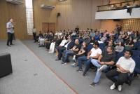 Foto mostra uma pessoa em pé, no palco de um auditório realizando uma palestra. E na mesma foto mostra outras pessoas sentadas acompanhando a palestra.