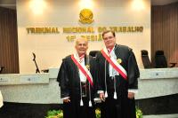 Foto mostra dois homens, dentro de um plenário. Ambos estão em pé e usam togas. Ao fundo, tem um brasão e o nome Tribunal Regional do Trabalho.