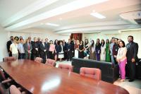 Foto mostra homens e mulheres em pé, posicionados no fundo de uma sala. No primeiro plano um tampo de uma grande mesa de madeira.