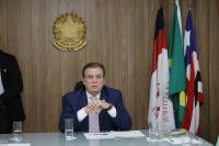 Foto de um homem branco usando um terno azul escuro. Ao lado direito dele as bandeiras do Brasil, Maranhão e da Justiça do Trabalho