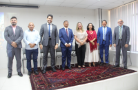 Foto de duas mulheres e seis homens lado a lado, de pé sobre um tapete vermelho estampado. Todos estão sorrindo para a câmera. Atrás deles, parede e persianas cinza claro.