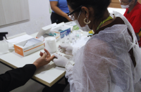 Foto mostra uma pessoa usando um jaleco branco. Ela está realizando um procedimento de teste rápido de coleta de sangue