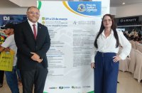 Foto mostra um homem e uma mulher em pé. Ambos estão ao lado de um painel de lona, mostrando a logomarca da Ação Praça de Justiça e Cidadania