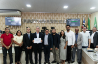 Foto mostra pessoas em pé, uma ao lado da outra. Ao centro um homem usando um terno, segura  um diploma. Ao fundo na parede: PLENÁRIO JOÃO FLOR DA SILVA.
