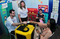 Foto mostra três pessoas. Duas delas estao sentadas. Uma outra está em pé ao lado de uma mesa amarela