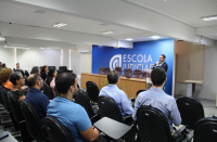 Foto mostra várias pessoas sentadas em um auditório. Ao fundo um homem, está em pé, diante do público.
