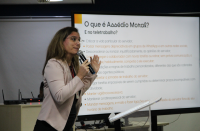 Foto mostra uma mulher em pé, segurando um microfone. Ao fundo um painel branco exibe a pergunta: O que é assédio moral?
