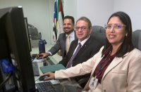 Foto mostra tres pessoas sentadas diante de um monitor de computador