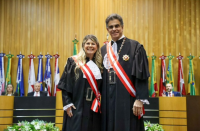 Foto mostra um homem e uma mulher. Ambos estão em pé. Ao fundo uma parede marron e várias bandeiras