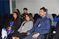 Foto mostra duas mulheres e um homem. Todos estão sentados em cadeiras dentro de um auditório