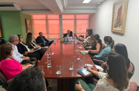 Foto mostra várias pessoas sentadas ao redor de uma mesa grande de reunião.