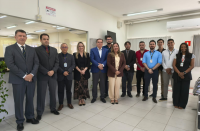Foto mostra um grupo formado por homens e mulheres em pé. Um ao lado do outro. Ambos estão em uma sala.