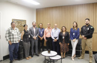 Foto mostra um grupo de pessoas em pé, dentro de uma sala. O grupo é formado por homens e mulheres.