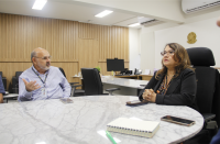 Foto mostra um homem e uma mulher. Ambos estão sentados juntos a uma mesa de reunião.
