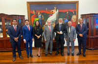 Foto mostra sete pessoas em pé, uma ao lado da outra. Ao fundo posicionado na parede um grande quadro com a imagem da Deus símbolo do Direito.