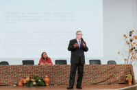 Foto de homem de pé, falando ao microfone, e mulher sentada atrás, à uma mesa.