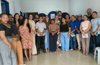 Foto mostra várias pessoas em pé, uma ao lado da outra dentro de uma sala. Ao fundo um pequeno cartaz com o nome PID.