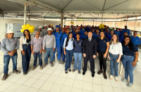 Foto mostra um grupo de pessoas em pé, olhando para a frente. Todos estão em um salão