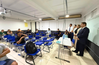Foto mostra uma sala de aula. Quatro pessoas estão em pé em frente as cadeiras e alguns universitários estão sentados.