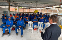 Foto mostra várioas pessoas sentadas em candeiras, acompanhando a palestra de um que está no primeiro plano da foto de frente para o público
