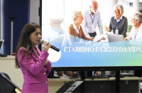 Foto mostra uma mulher usando um blaser na cor rosa. Ao fundo um telão projeto uma imagem com idosos reunidos. E a mensagem etarismo e ciclo da vida