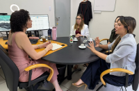 Foto mostra 4 mulheres sentadas em volta de uma mesa. Elas estão em uma sala, ao fundo dois monitores de computador.