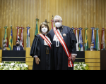 Imagem de parte do Plenário, com fundo em madeira, onde estão alinhadas várias bandeiras, dois homens sentados na bancada e ao centro a ministra Maria Cristina Peduzzi e o ministro Emmanoel Pereira 