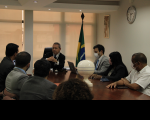Fundo branco com imagem do presidente do TRT, desembargador Carvalho Neto, trajando terno escuro, camisa branca, gravata escura, estando sentado ao lado do procurador do trabalho Maurício Pessoa Lima, vestido com terno escuro. Ambos estão de frente para os demais participantes da audiência que se encontram sentados em torno da mesa de reunião.