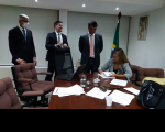 Foto da desembargadora Márcia Andrea, sentada à mesa de reunião no Gabinete da Presidência, vestindo blazer cinza, usando óculos, assinando ata de audiência de conciliação, observada pelo procurador do trabalho Roberto Moreira, pelo assessor do gabinete da desembargadora, Hermano Rocha Filho, e o procurador do município Bruno Duailibe, os três em pé, com ternos escuros