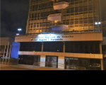 Foto da fachada do prédio-sede do TRT no Maranhão, no bairro Areinha. A fachada está iluminada na cor amarela em adesão ao Maio Amarelo, e se vê partes do projeto arquitetônico nas cores azul e bege, uma pequena grande antes do acesso à Portaria do Tribunal (frontal), na lateral esquerda os mastros de hastear as bandeiras oficiais