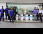 Foto de servidores, servidoras, terceirizadas e terceirizados em um dos ambientes do prédio-sede do Tribunal do Trabalho onde se vê de coletores de papel, plástico, metal e lixo comum, ao fundo uma parede de vidro e parte de parede pintada na cor branca